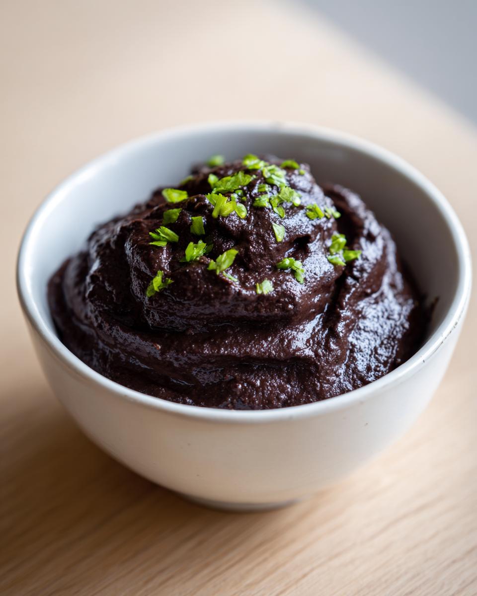 A close-up of rich, dark black bean dip topped with chopped green herbs in a white bowl.