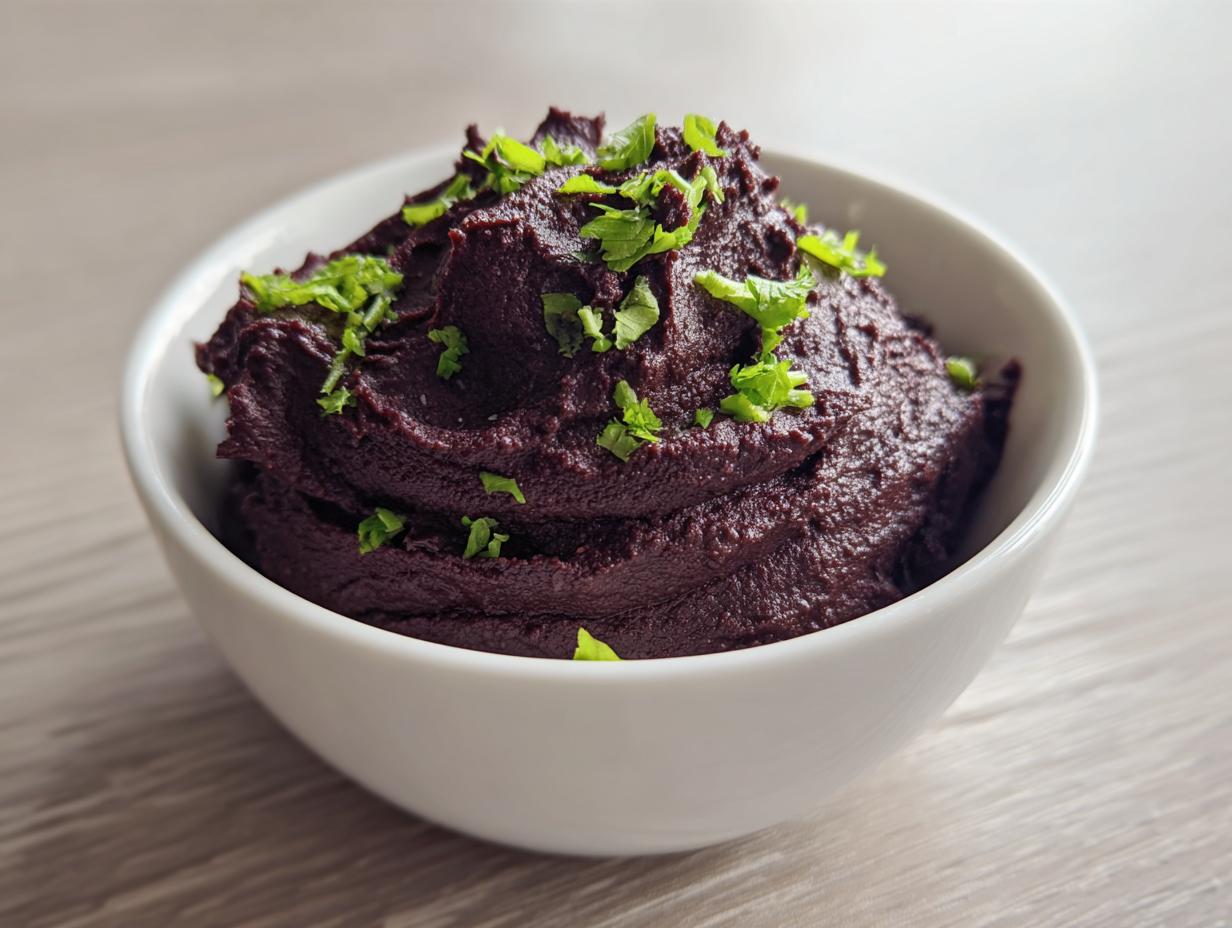 A close-up of rich, dark purple-brown black bean dip mounded high in a white bowl and topped with fresh green herbs.