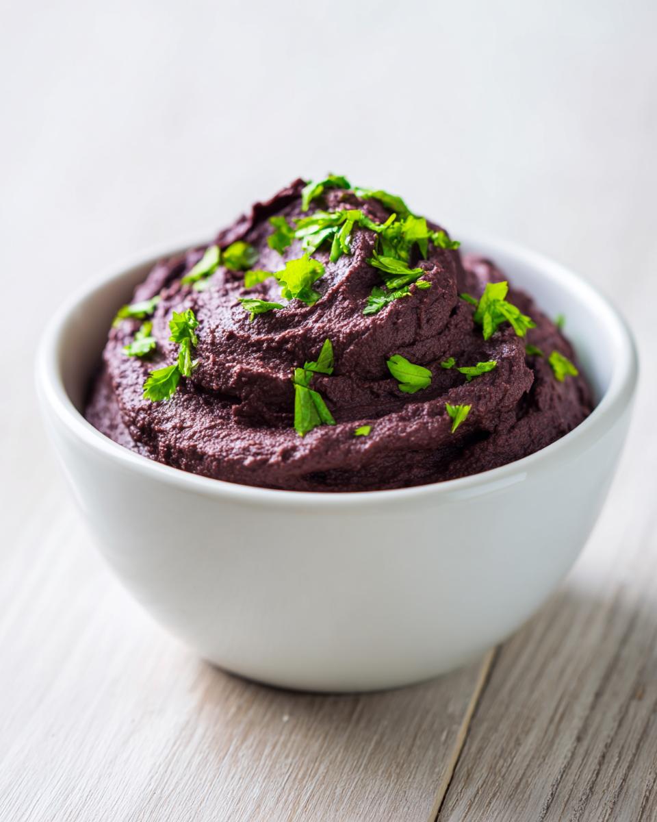 A close-up of rich, dark purple black bean dip mounded in a white bowl and topped with fresh green parsley.