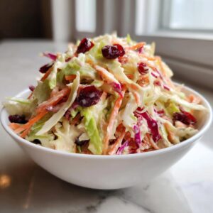 A close-up of creamy apple cranberry coleslaw, featuring shredded cabbage, carrots, and dried cranberries, served in a white bowl.