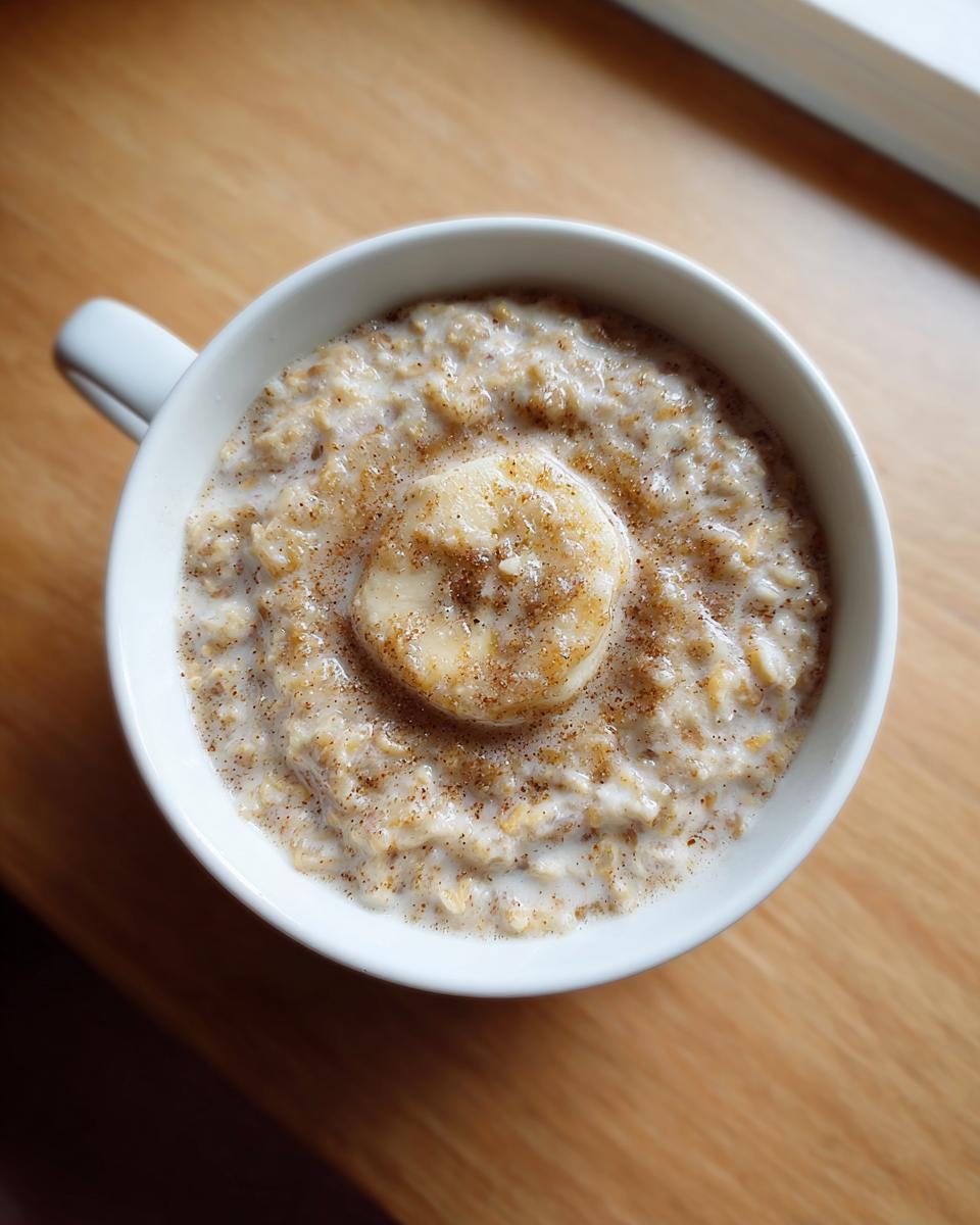 Overhead view of creamy banana oatmeal topped with a slice of banana and cinnamon in a white mug.