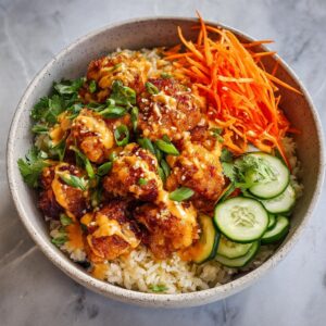 A close-up of a Bang Bang Salmon Bowl featuring crispy salmon bites over rice, topped with creamy sauce, shredded carrots, cucumber slices, and cilantro.