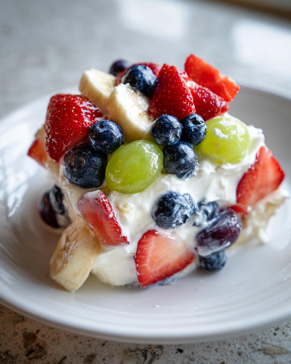 A serving of creamy cheesecake fruit salad topped generously with strawberries, blueberries, bananas, and green grapes.