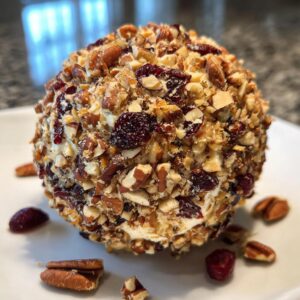 A festive cranberry pecan cheese ball coated in chopped nuts and dried cranberries, served on a white plate with scattered pecans and cranberries.