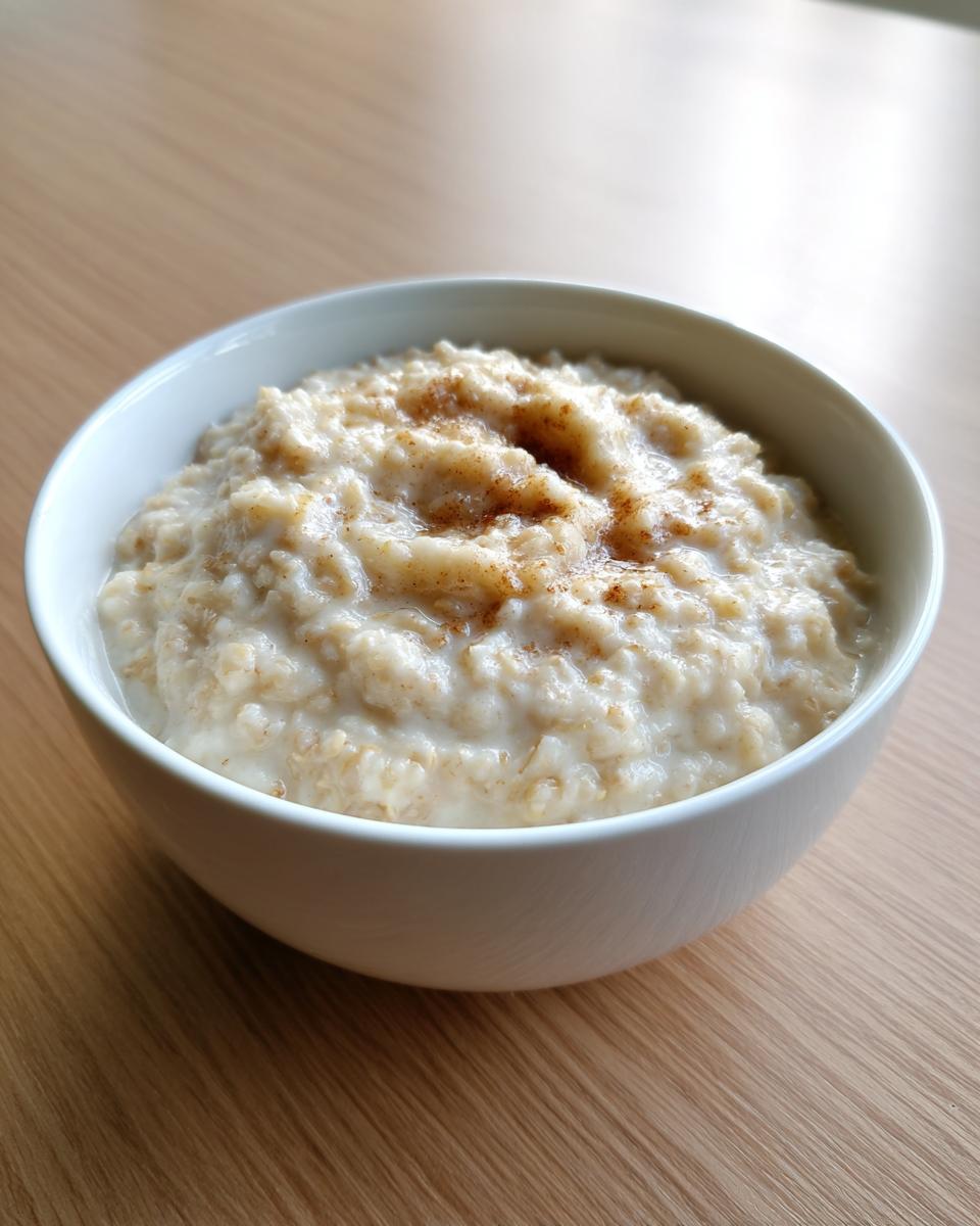 A close-up of a white bowl filled with creamy banana oatmeal, lightly sprinkled with cinnamon.