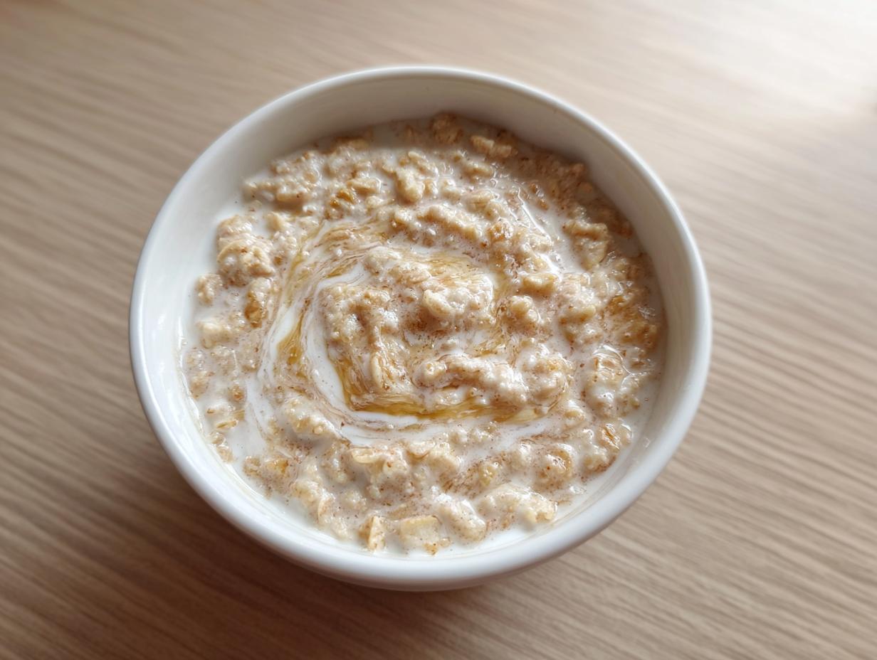 Overhead view of a white bowl filled with creamy banana oatmeal, topped with milk and a swirl of honey.