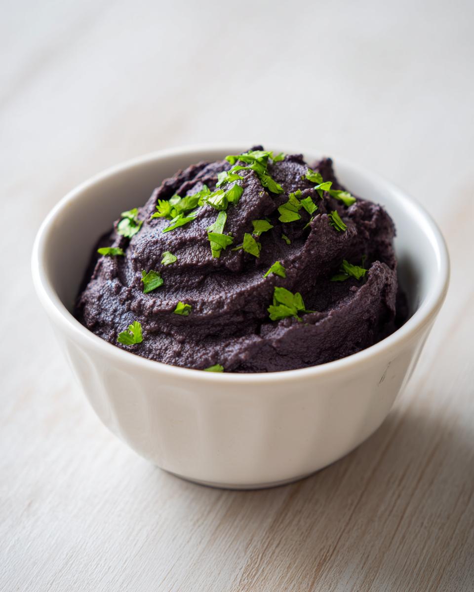A close-up of rich, dark purple black bean dip topped with fresh chopped parsley in a white bowl.