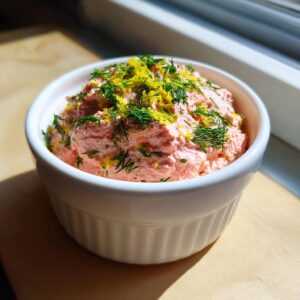 A creamy, pink salmon dip topped with fresh dill and bright yellow lemon zest in a white bowl.
