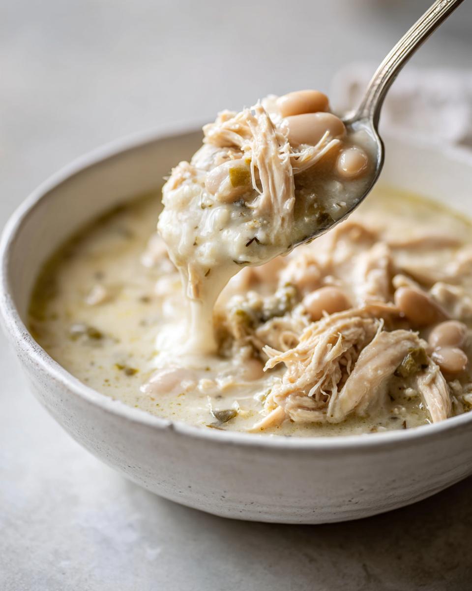 A spoonful of creamy white chicken chili, featuring shredded chicken and white beans, being lifted from a bowl.
