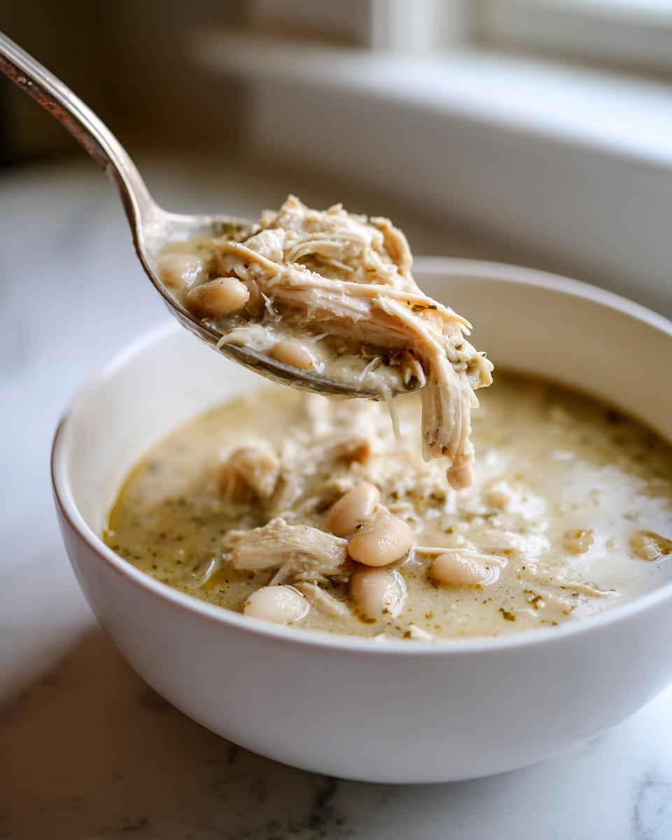 A spoonful of creamy white chicken chili, showing shredded chicken and white beans, lifted from a bowl.