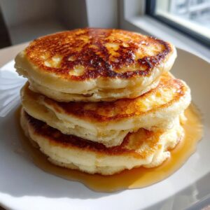 A stack of three fluffy ricotta pancakes drizzled with maple syrup on a white plate.