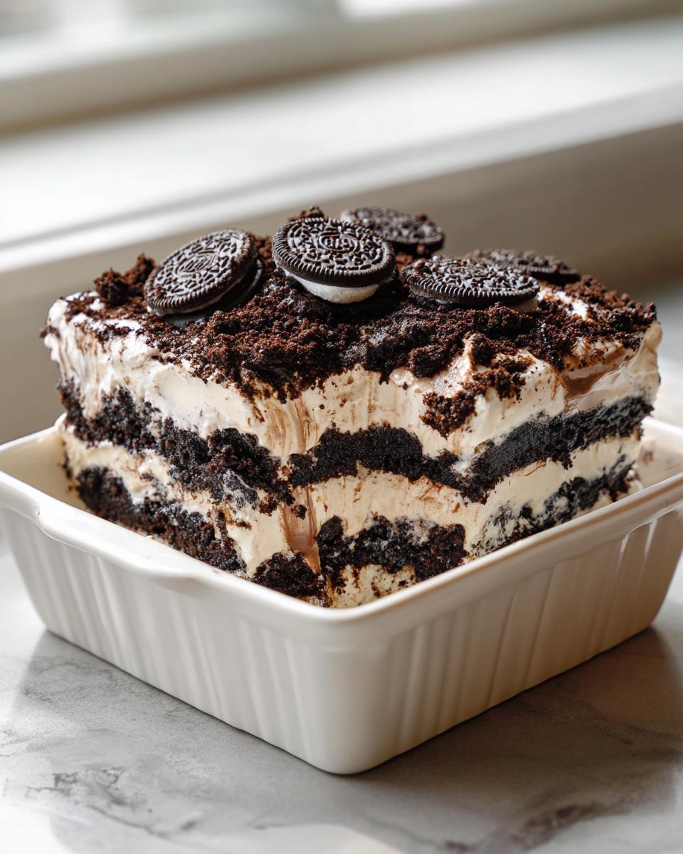 A square slice of layered dirt cake featuring alternating layers of chocolate cookie crumbs and cream, topped with whole Oreos.