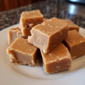 A pile of perfectly cut squares of creamy peanut butter fudge on a white plate.