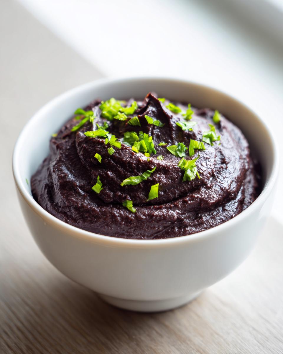 A close-up of thick, dark purple black bean dip garnished with bright green chopped chives in a white bowl.