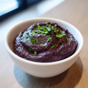 A close-up of thick, rich purple black bean dip served in a white bowl and topped with fresh chopped parsley.