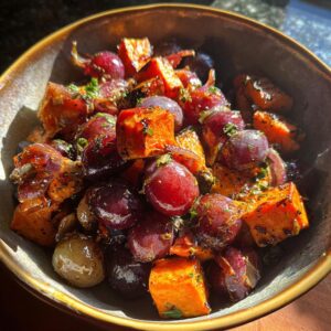 Close-up of a bowl filled with glistening roasted grapes and sweet potatoes, seasoned with herbs.