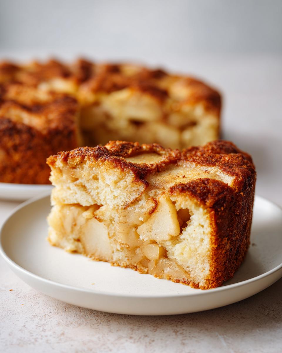 A thick slice of moist apple cake, packed with soft apple pieces and a cinnamon sugar top, served on a small white plate.