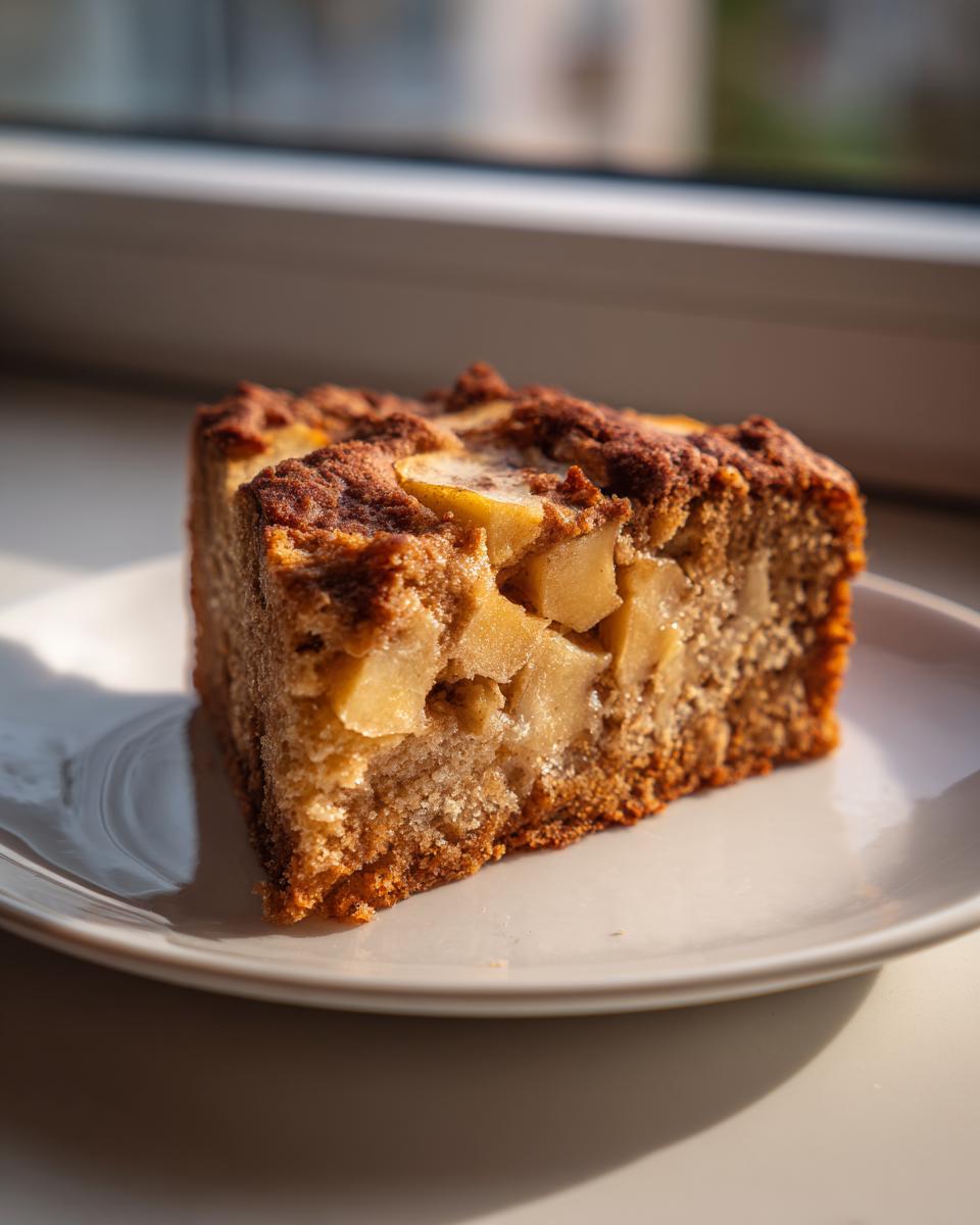 A close-up slice of moist apple cake, rich in cinnamon flavor and loaded with visible apple chunks, resting on a white plate.