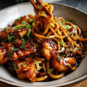 Close-up of chopsticks lifting glossy sticky garlic chicken noodles from a bowl, garnished with green onions.