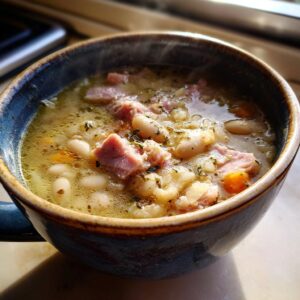 A steaming bowl of white bean and ham soup, filled with tender beans, chunks of ham, and visible herbs.