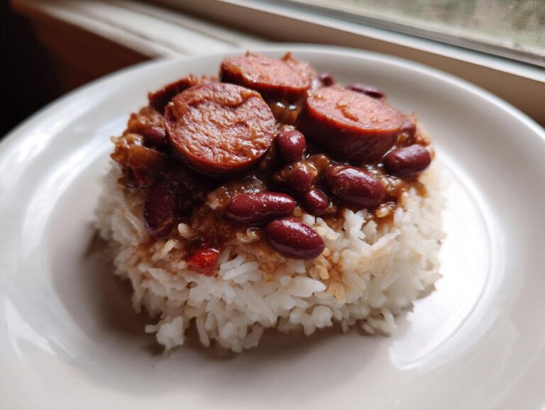 A serving of amazing red beans and rice topped with sliced smoked sausage on a white plate.