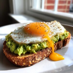 Delicious avocado toast topped with a sunny-side-up egg, featuring runny yolk dripping down the side.