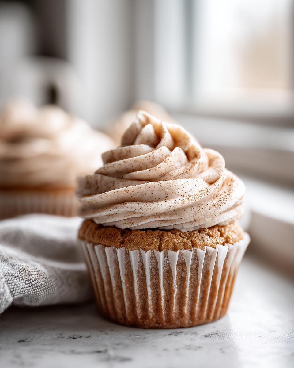 A beautifully frosted banana cupcake topped with cinnamon spice, sitting next to a linen napkin.