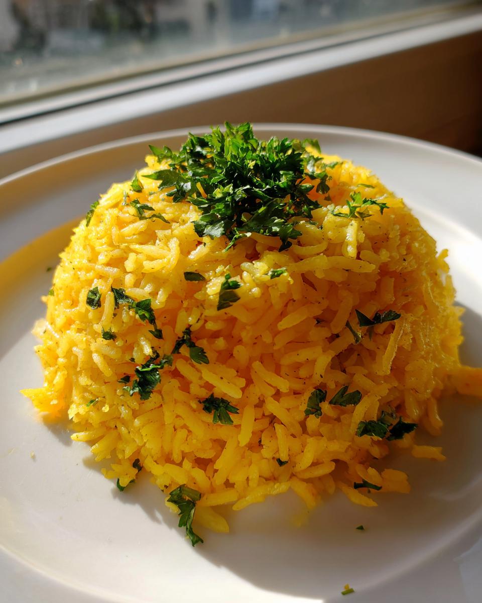 A mound of bright yellow mediterranean rice topped with fresh chopped parsley, served on a white plate near a window.
