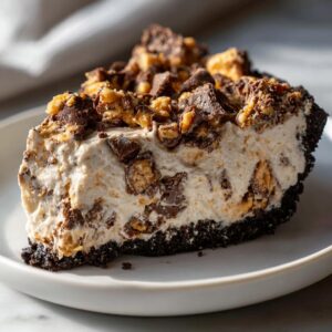 A generous slice of creamy no-bake butterfinger pie with a dark chocolate crust and topped with crushed candy pieces.