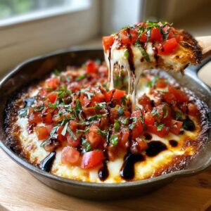A scoop of hot, cheesy bruschetta dip is pulled from a cast iron skillet, showing a long cheese stretch.