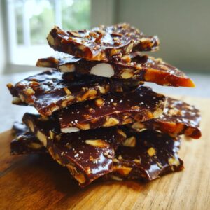 Stack of dark chocolate almond toffee pieces sprinkled with sea salt on a wooden board.