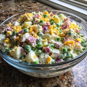 Close-up of a vibrant, creamy pea salad mixed with ham, eggs, and red onion in a clear glass bowl.
