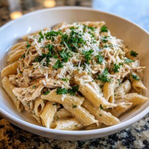 A close-up of a white bowl filled with creamy crockpot garlic parmesan chicken pasta, topped with shredded chicken, cheese, and parsley.