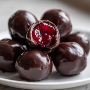 A pile of glossy, dark chocolate covered cherries, with one piece broken open showing the bright red cherry filling.