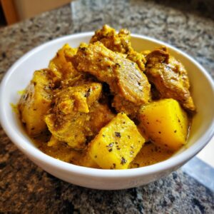 A close-up of rich, yellow Jamaican curry chicken pieces mixed with chunks of potato in a white bowl.