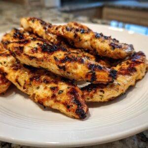 Close-up of perfectly grilled, juicy greek chicken tenders seasoned with herbs on a white plate.