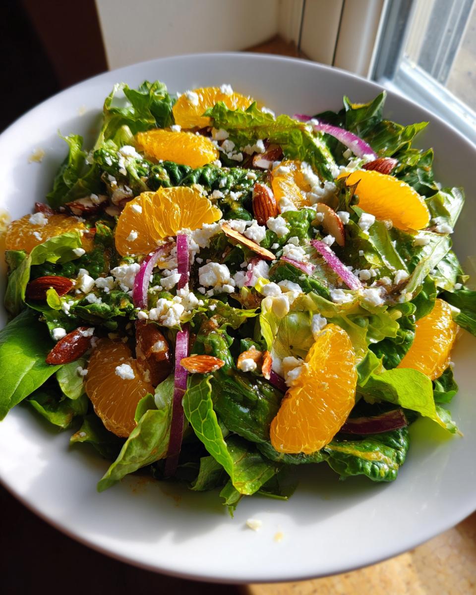 A bright bowl of mandarin orange salad featuring crisp greens, orange slices, feta cheese, almonds, and red onion.
