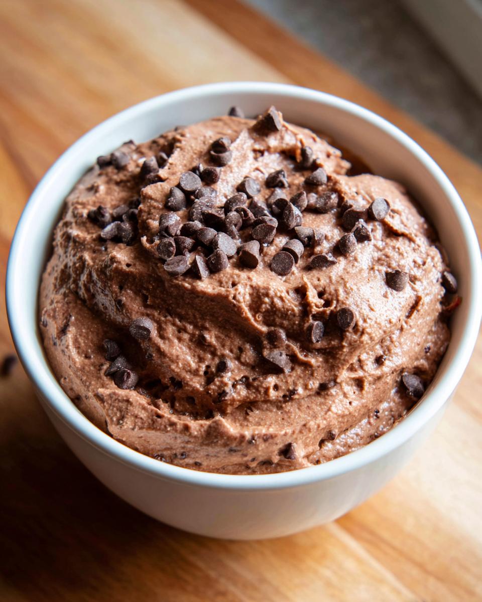 A close-up of rich, thick brownie batter dip in a white bowl, generously topped with mini chocolate chips.