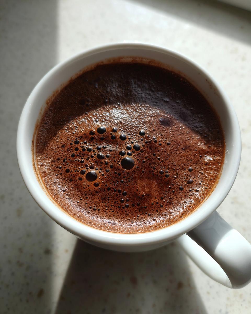 Overhead close-up of rich, dark brown hot chocolate with small bubbles on top in a white mug.