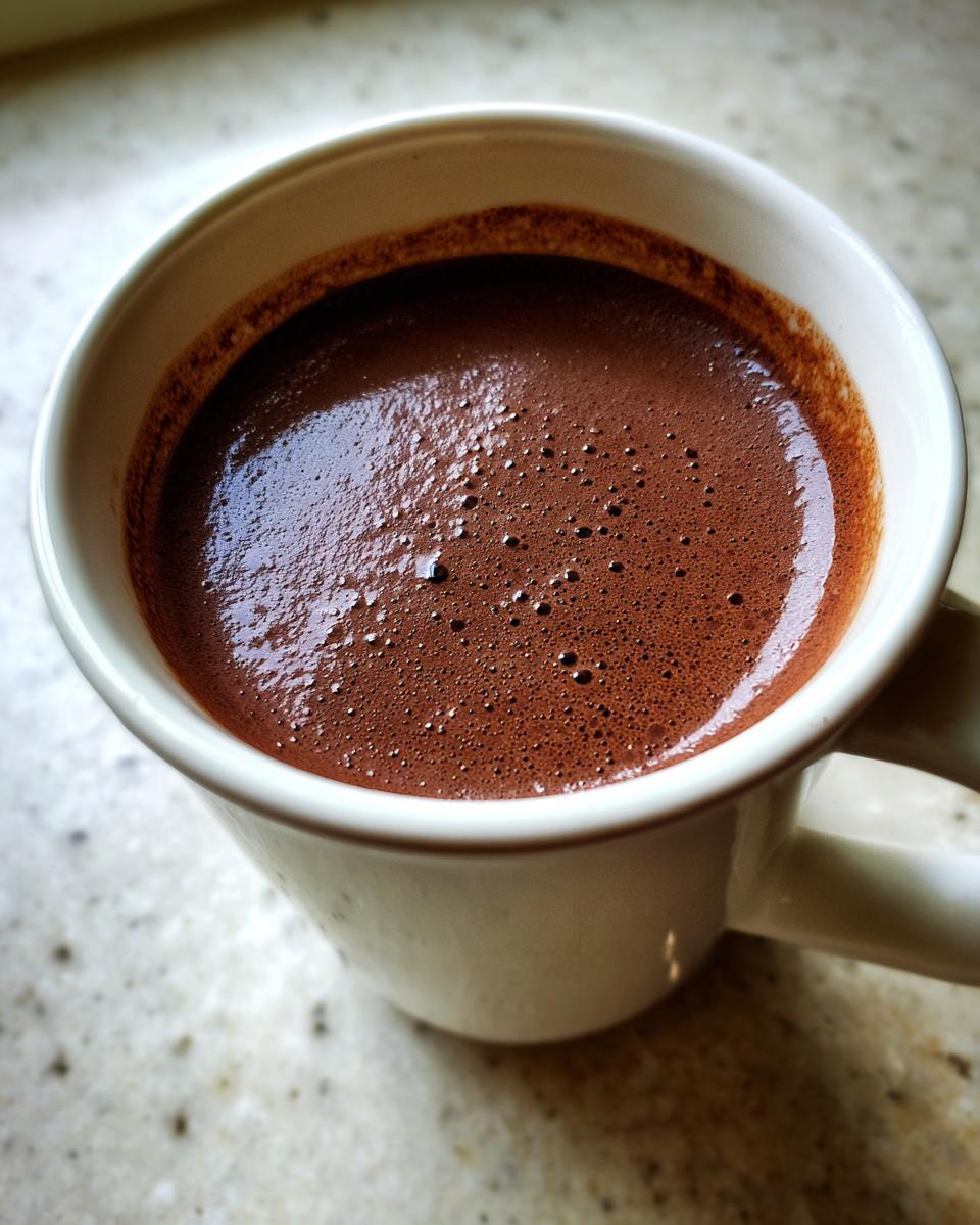 Close-up of rich, dark brown hot chocolate with a layer of small bubbles in a white ceramic mug.