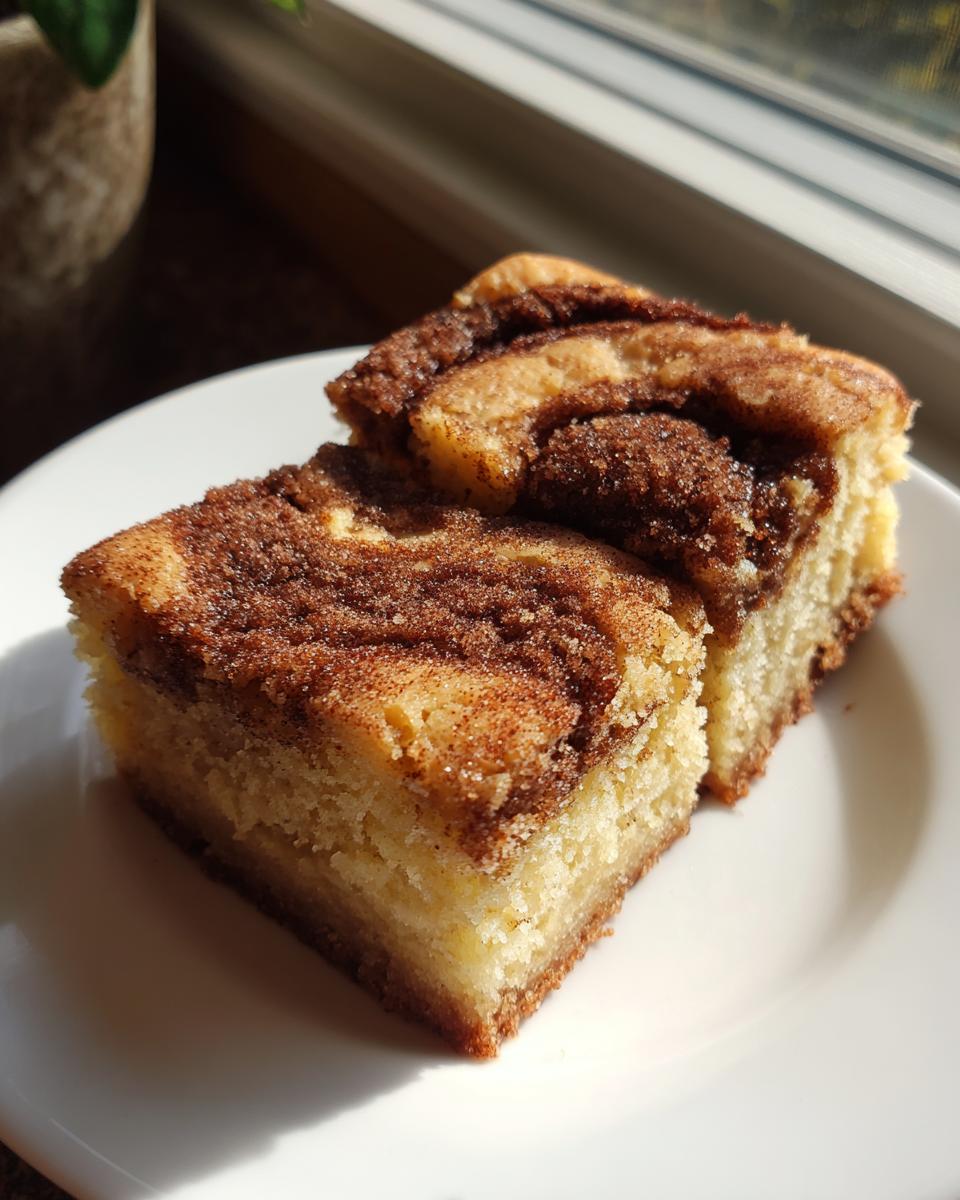 Two square slices of rich snickerdoodle blondies topped with a thick, sugary cinnamon swirl, served on a white plate.