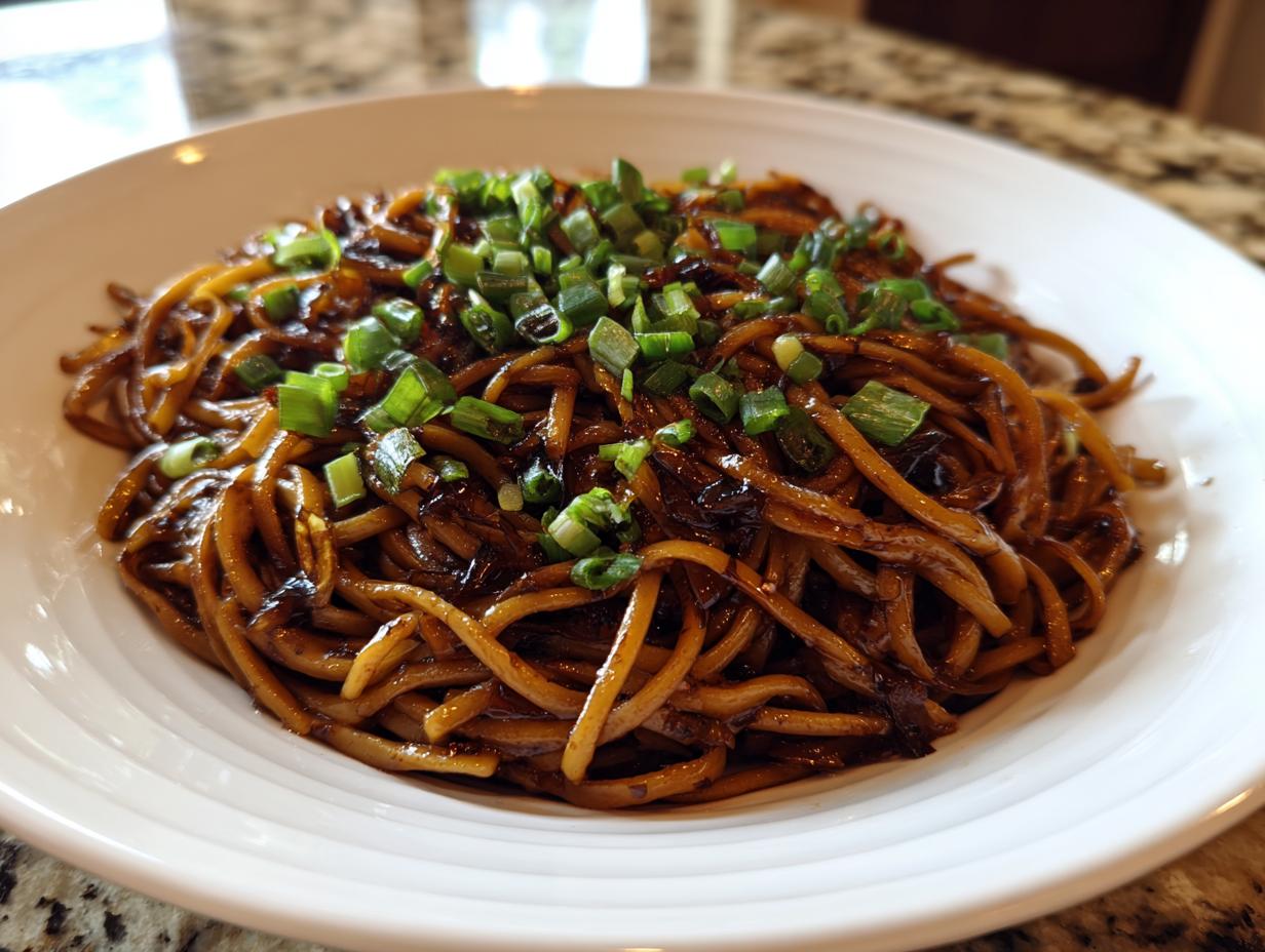 A close-up shot of a bowl filled with glossy, dark-sauced garlic noodles topped with fresh chopped green onions.