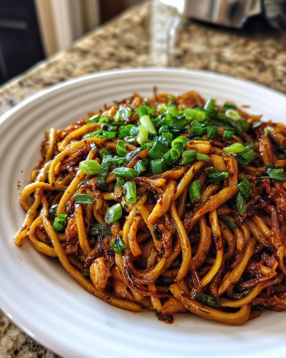 A close-up of a white plate piled high with saucy, thick garlic noodles topped generously with fresh chopped green onions.