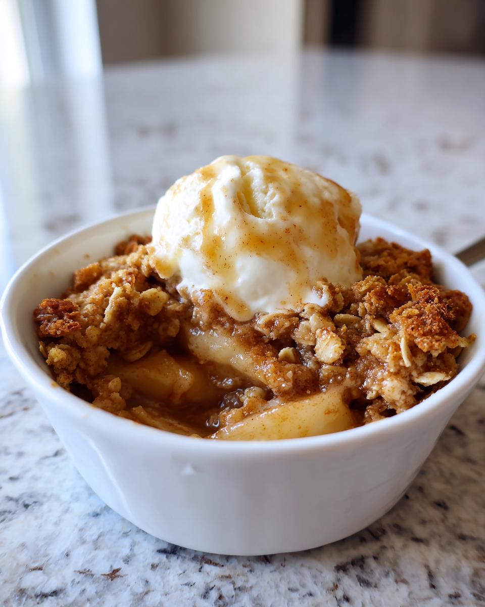 Close-up of a warm apple crisp recipe served in a white bowl, topped with vanilla ice cream and caramel drizzle.