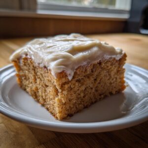 A moist slice of applesauce cake topped with thick, creamy frosting on a white plate.