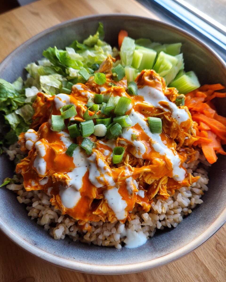 A close-up of a healthy buffalo chicken bowl featuring shredded buffalo chicken over brown rice, topped with ranch dressing and green onions.