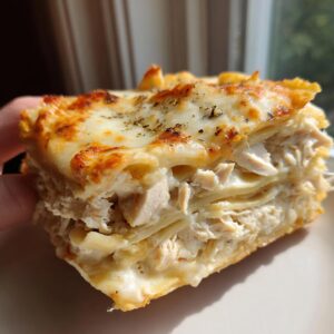A thick, layered slice of cheesy chicken alfredo lasagna held up close, showing shredded chicken filling.