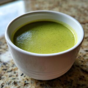 Close-up of creamy, vibrant green celery soup served in a white bowl on a speckled countertop.