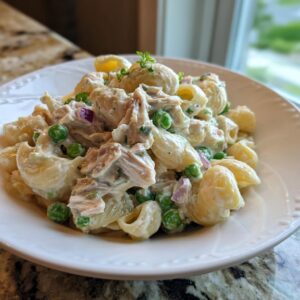 A close-up of creamy Tuna pasta salad mixed with shell pasta, green peas, and red onion, served on a white plate.