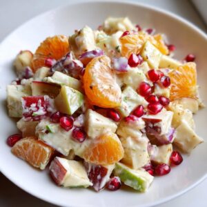 Close-up of a creamy winter fruit salad featuring diced apples, mandarin orange segments, and bright red pomegranate seeds.